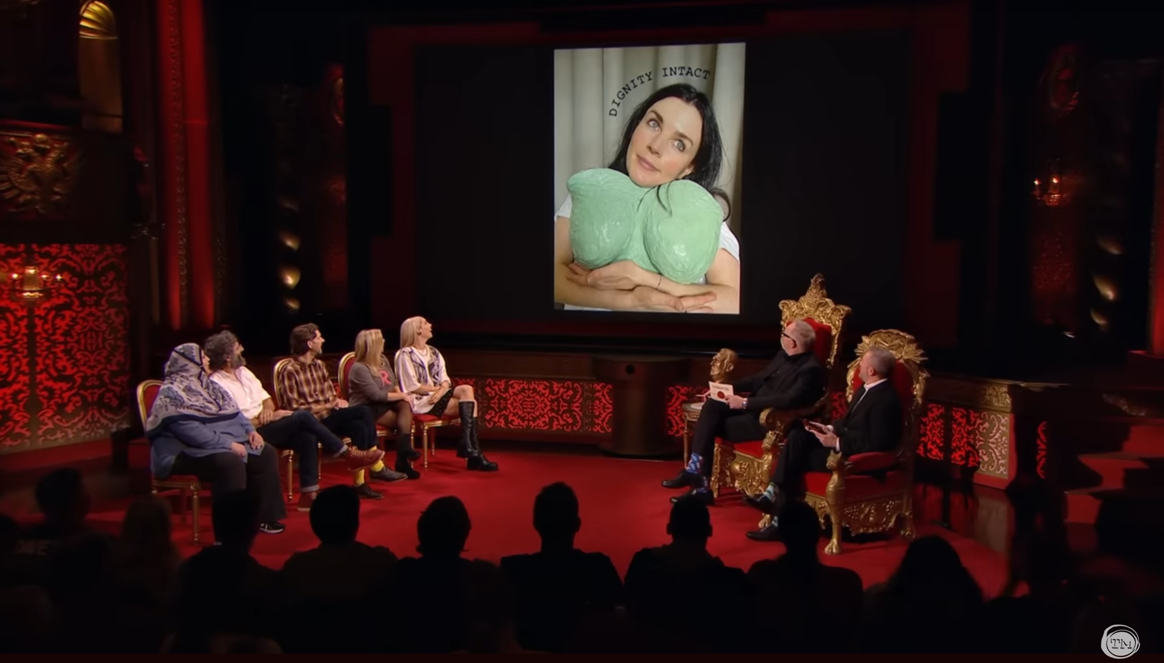 An in-studio shot of all the Taskmaster contestants looking at a photo of Aisling Bea, who is cradling a mold of her own breasts. The picture is captioned, “Dignity intact.”