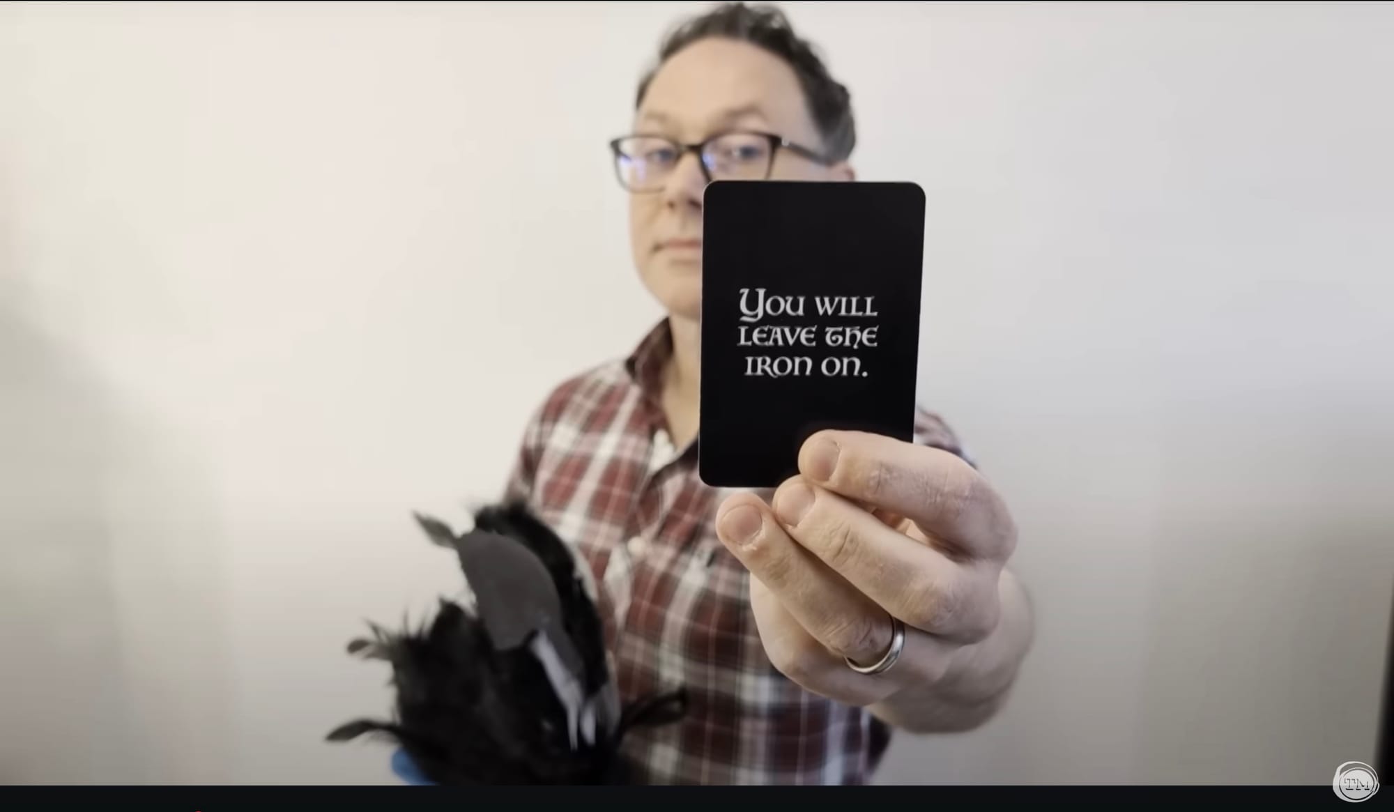Reece Shearsmith holds a fake raven in one hand and shows a fortune-telling card that says, “You will leave the iron on.‘