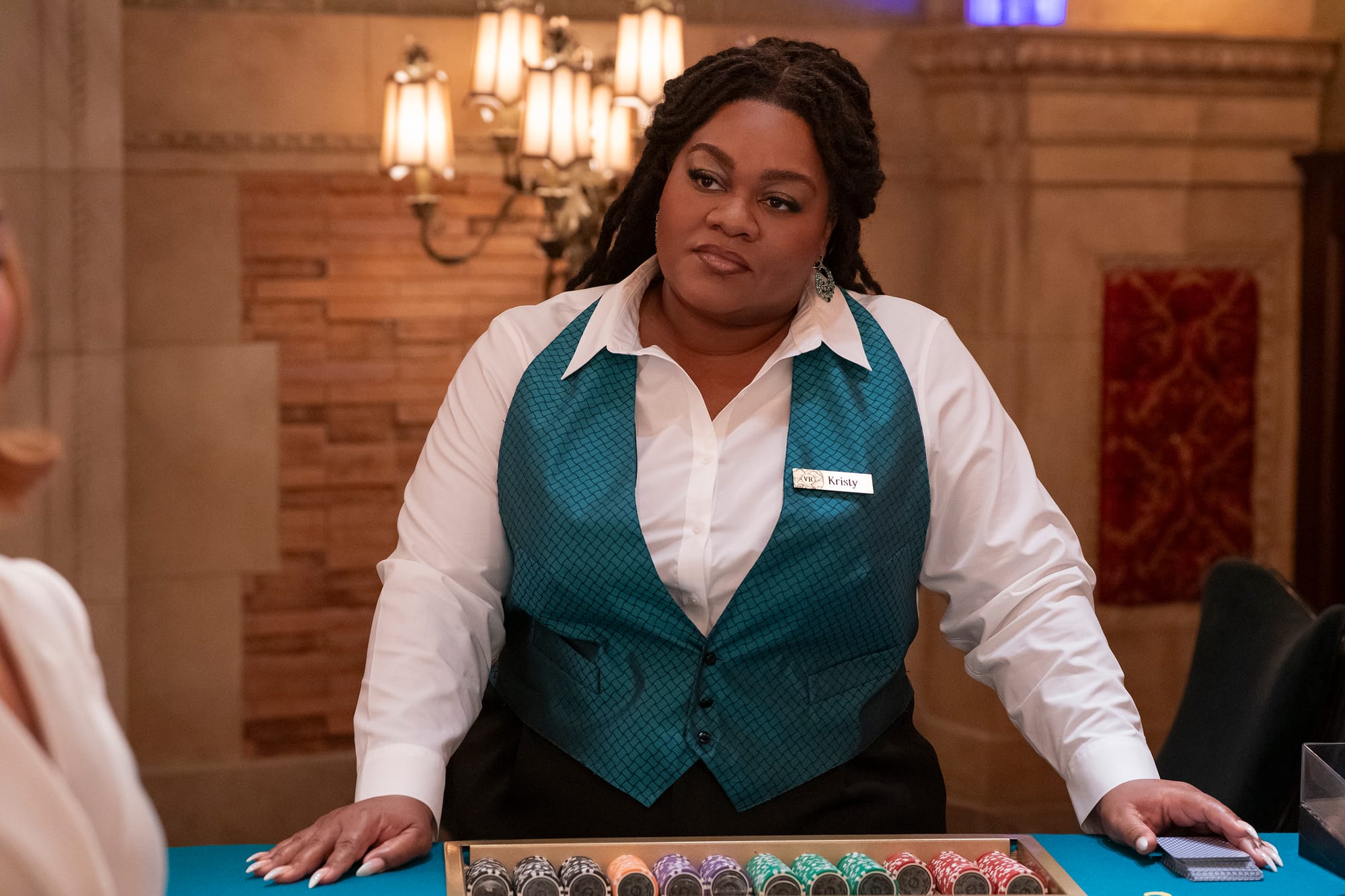 Da'Vine Joy Randolph wears a turquoise vest and a white dress shirt. She stands behind a rack of gambling chips.