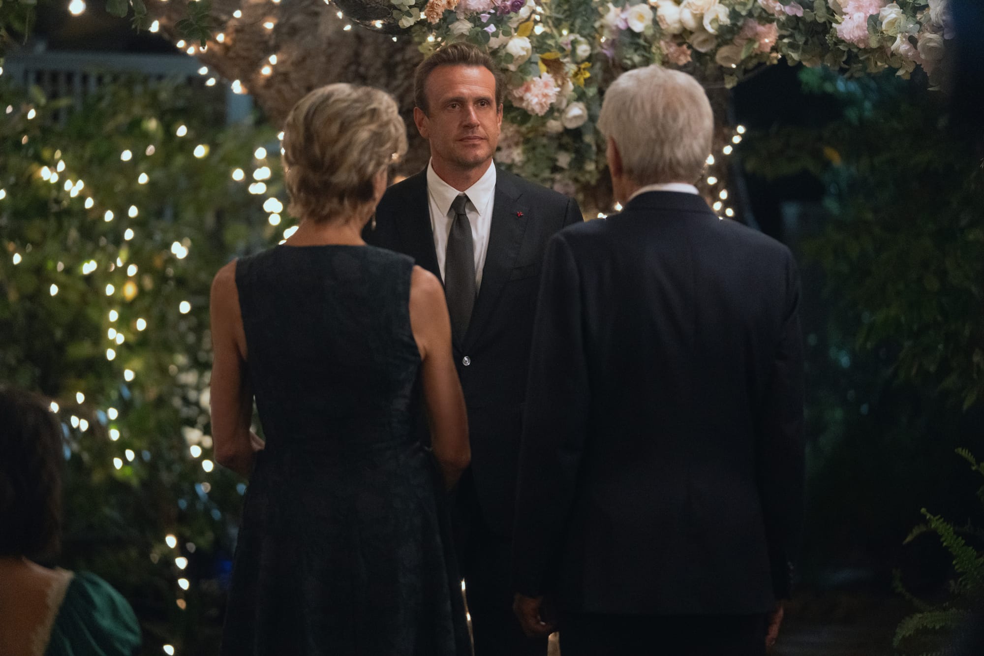 Jason Segel stands in front of a tree decorated for a wedding. Wendie Mallick and Harrison Ford have their backs to the camera.