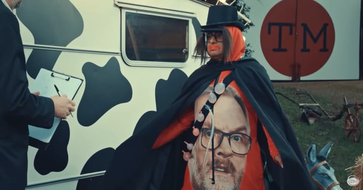 Sanjeev Bhaskar dressed in a hat, wig, and a onesie with Greg Davies’ face printed on it.