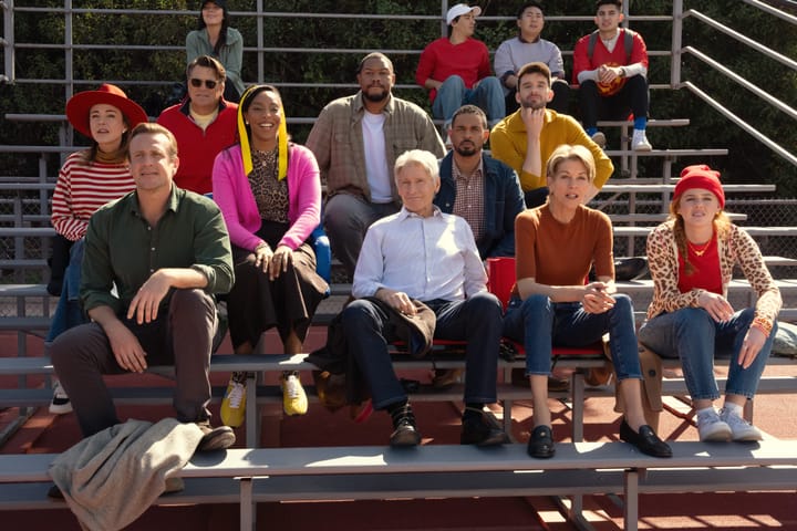 Almost the entire cast of Shrinking on a set of metal bleachers, watching a high school soccer game.