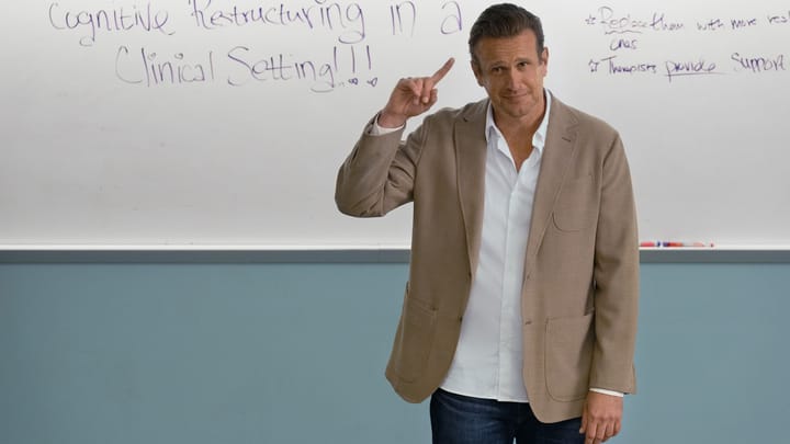 Jason Segel stands in front a whiteboard in a lecture hall, pointing to his head